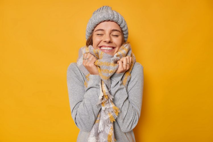 Portrait of happy young woman wears warm clothes knitted hat scarf around neck closes eyes with satisfaction smiles gladfully with white teeth isolated over yellow background has pleasant memory