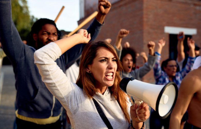 Young displeased woman using megaphone and shouting while protesting with group of people on city streets.