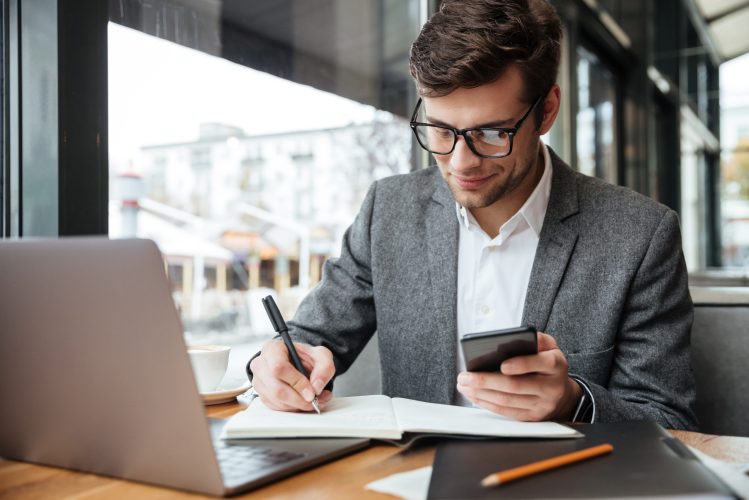 Smiling business man in eyeglasses sitting by the table in cafe with laptop computer while using smartphone and writing something