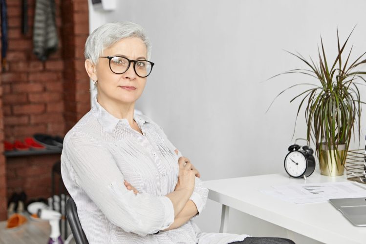 Age, maturity, profession and employment. Indoor shot of confide Age, maturity, profession and employment. Indoor shot of confident serious fifty year old female freelancer working at home office while being on retirement, using laptop, crossing arms on her chest