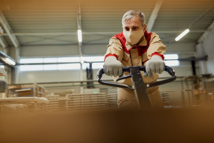 Mid adult carpenter pulling pallet jack and wearing protective face mask in a workshop due to coronavirus pandemic.