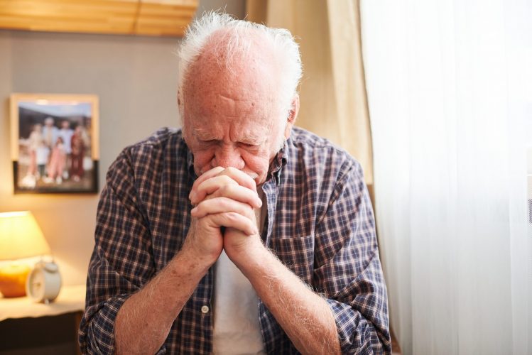 Senior man with white hair looking very sad while sitting on the armchair and thinking about something in his room