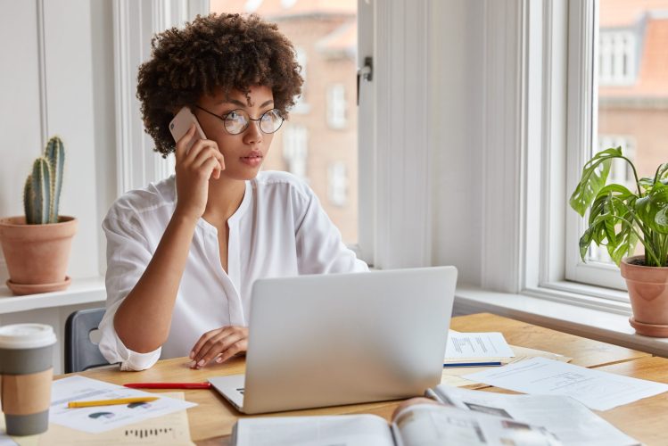 Serious ethnic female CEO wears round spectacles, tries to reach decision with colleague via cell phone, uses laptop computer, surrounded with papers, studies graphs, notices decrease in prices