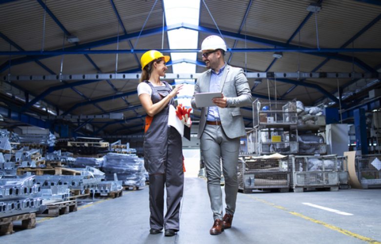 Manager supervisor and industrial worker in uniform walking in large metal factory hall and talking about increasing production.