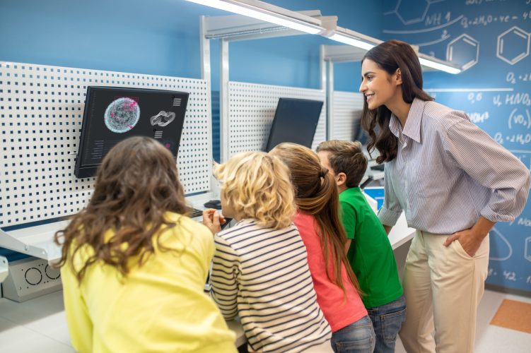 Information. Teacher sideways to camera and children standing with their backs looking at screen hanging on wall in classroom