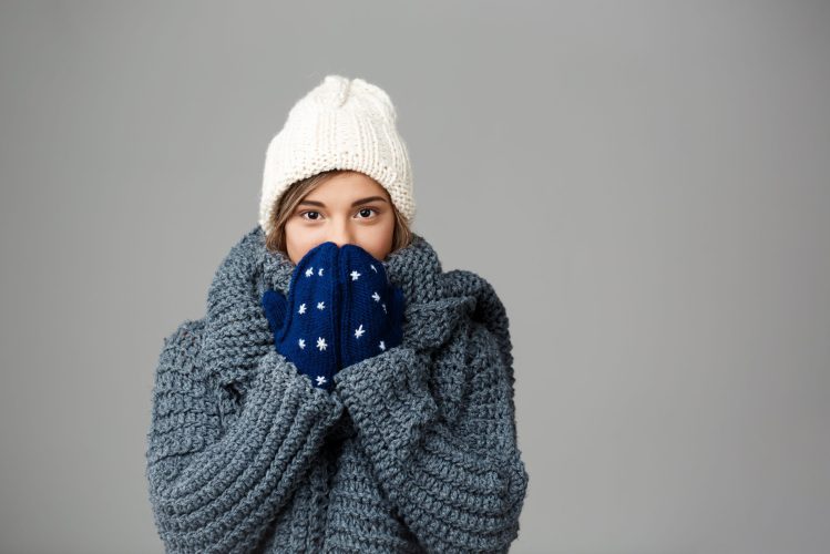 Young beautiful fair-haired girl in knited hat sweater and mittens smiling looking at camera over grey background. Copy space.